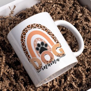 May include: White ceramic coffee mug with a rainbow design featuring a leopard print pattern. The rainbow is in shades of orange and brown. The text "DOG mama" is written below the rainbow.