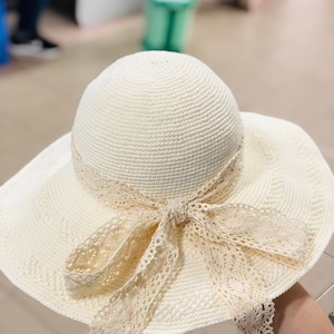 Peut inclure: Un chapeau de soleil crème avec un large bord et une couronne crochetée. Le chapeau est orné d'un délicat ruban en dentelle noué en nœud autour de la base de la couronne. Le bord est également crocheté.