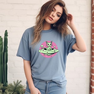 May include: A light blue t-shirt featuring a pink circle graphic of a green cat sitting in a flying saucer. The cat has large, pink eyes and a white star above its head.