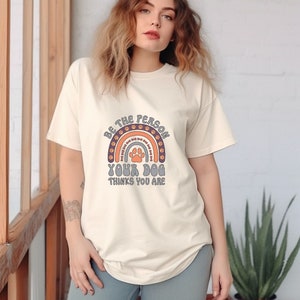 May include: A cream colored t-shirt with a rainbow design featuring paw prints. The text on the shirt reads "Be the person your dog thinks you are."