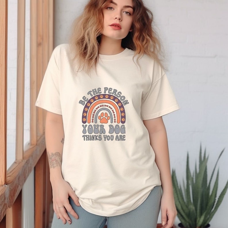 May include: A cream colored t-shirt with a rainbow design featuring paw prints. The text on the shirt reads "Be the person your dog thinks you are."