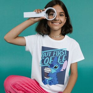 以下が含まれることがあります： コーヒーカップを持った青い漫画のキャラクターが描かれた白いTシャツ。Tシャツには「But First Coffee」という文字が印刷されています。