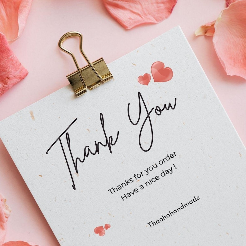 May include: A white card with the words "Thank You" in elegant script, topped with a gold binder clip. The card features two red heart illustrations and the text "Thanks for your order, Have a nice day!" on a pink background.