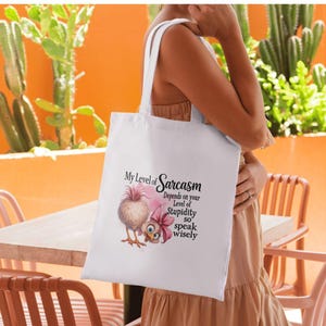 May include: White tote bag with the text "My Level of Sarcasm Depends on your Level of Stupidity so speak wisely" and a cartoon bird illustration. The bag has two white straps and is held by a person.