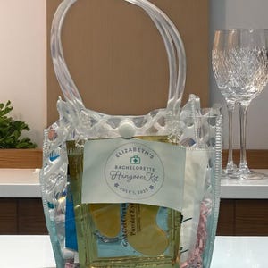 May include: Clear plastic tote bag filled with hangover recovery items. The bag has a clear handle and a white tag that reads "Elizabeth's Bachelorette Hangover Kit" with the date July 1, 2025. Gold gel eye masks and other items are visible inside.