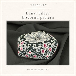 May include: A handcrafted biscornu pincushion featuring the Lunar Silver biscornu pattern. The design displays intricate cross-stitch embroidery in shades of black, silver, and magenta, with a central silver bead. Text reads TREASURY and ATELIER PIN & NEEDLE.