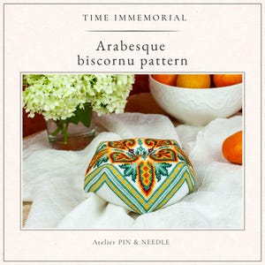 Op de afbeelding: Een biscornu-patroon met een arabesk-ontwerp, in groen, geel, rood en wit. De tekst "TIME IMMEMORIAL" en "Arabesque biscornu pattern" staan bovenaan. De tekst "Atelier PIN & NEEDLE" staat onderaan. Het ontwerp staat op een witte achtergrond.