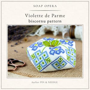 Może przedstawiać: Biała poduszeczka na szpilki z tkaniny z wyhaftowanym ściegiem krzyżykowym wzorem biscornu Violette de Parme w odcieniach niebieskiego i zielonego. Tekst SOAP OPERA, Violette de Parme biscornu pattern oraz ATELIER PIN & NEEDLE jest nadrukowany.