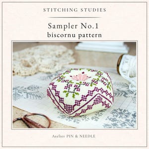Puede incluir: Un cojín cuadrado de color blanquecino con un diseño floral de punto de cruz en tonos rosa, verde y morado. El cojín está etiquetado como "Sampler No.1 biscornu pattern" y está rodeado de artículos de costura. También se ve el texto "Stitching Studies" y "Atelier PIN & NEEDLE".