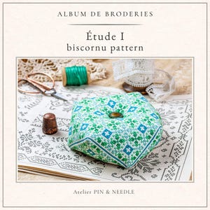May include: A square, green and blue biscornu pattern with a button in the center, displayed on a vintage book. The image includes a thimble, scissors, thread, and lace. Text reads "Album de Broderies", "Étude I biscornu pattern", and "Atelier PIN & NEEDLE".