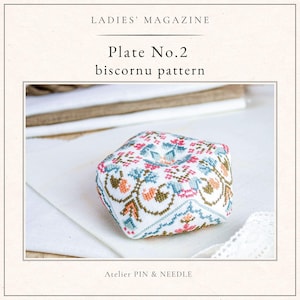 Peut inclure: Un coussin à épingles carré avec un motif floral au point de croix rose, bleu et orange. Le texte "LADIES' MAGAZINE" et "Plate No.2 biscornu pattern" sont visibles. Les mots "Atelier PIN & NEEDLE" sont également présents.