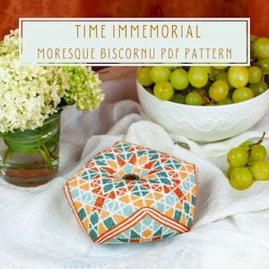 Op de afbeelding: Een kleurrijk, geometrisch gepatroneerd, zeshoekig naaldwerk kussen met een knoop in het midden. Het kussen ligt op een wit stofoppervlak met een witte kom groene druiven en een vaas met witte bloemen op de achtergrond. De tekst "TIME IMMEMORIAL MORESQUE BISCORNU PDF PATTERN" staat in een lichtbruine rechthoek bovenaan de afbeelding.