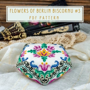 May include: A white fabric biscornu with a colorful floral design. The biscornu is a type of decorative pillow or pincushion. The design features pink, yellow, green, and blue flowers. The text "FLOWERS OF BERLIN BISCORNU #3 PDF PATTERN" is displayed above the biscornu.