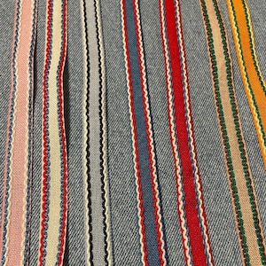 May include: A close-up of a variety of colourful ribbon trims with scalloped edges. The trims are arranged in rows on a blue denim fabric background.