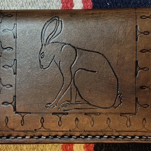 May include: A brown leather wallet with a detailed embossed image of a hare. The wallet features decorative border elements and is stitched with white thread. The hare is centered within a rectangular frame.