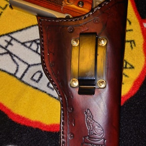 Könnte beinhalten: Ein braunes Lederholster mit einem Gürtelclip und einem Wolfdesign. Das Holster ist handgefertigt in den USA.