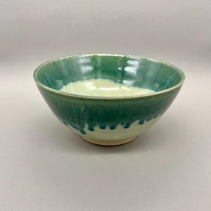 May include: A handmade ceramic bowl with a glossy green glaze. The bowl features a gradient of colors, transitioning from a light green interior to a darker green exterior. The bowl has a round shape and is suitable for serving food.