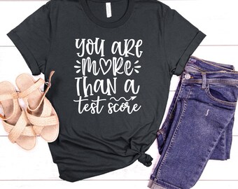 Könnte beinhalten: Ein dunkelgraues T-Shirt mit dem Text "You are more than a test score" in Weiß. Der Text enthält ein Herzsymbol und einen Pfeil.