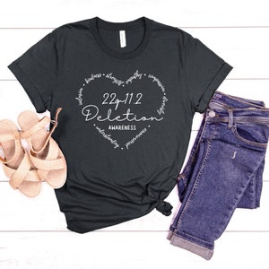 May include: A dark gray t-shirt with a white heart design. The heart has the text "22q11.2 Deletion Awareness" inside it. The words "kindness, strength, empathy, compassion, resilience, understanding, perseverance, hope, and happy" are written around the heart.