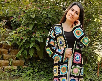 Könnte beinhalten: Ein langer schwarzer Häkelcardigan mit bunten quadratischen Mustern. Der Cardigan hat einen V-Ausschnitt und lange Ärmel.