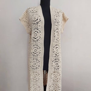 Könnte beinhalten: Ein langer, cremefarbener Häkelcardigan mit V-Ausschnitt und kurzen Ärmeln. Der Cardigan ist aus einem zarten, durchbrochenen Muster gearbeitet.