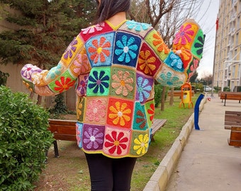 Könnte beinhalten: Eine bunte gehäkelte Strickjacke mit Blumenmuster. Die Strickjacke besteht aus Quadraten mit verschiedenen farbigen Blumen. Die Strickjacke wird von einer Person getragen, die auf einem Bürgersteig steht.