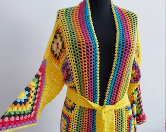 Könnte beinhalten: Ein langer, gelber Häkelcardigan mit einem Regenbogenstreifenmuster und einem Bindegürtel. Der Cardigan ist aus verschiedenen bunten Garnen gefertigt und hat eine lockere, fließende Passform.