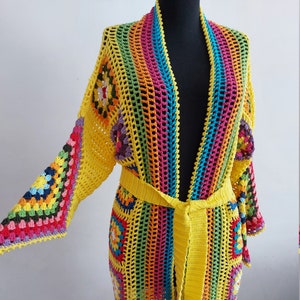 Può includere: Un lungo cardigan in crochet giallo con un motivo a righe arcobaleno e una cintura da annodare. Il cardigan è realizzato con una varietà di filati colorati e ha una vestibilità ampia e fluida.