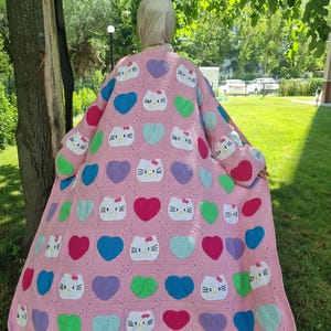 May include: A long, pink crochet cardigan featuring a repeating pattern of white Hello Kitty faces with red bows and multi-colored hearts in shades of blue, green, purple, and magenta, constructed from square panels with long sleeves.