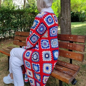 May include: A woman wearing a long, crocheted cardigan with a colorful geometric pattern in shades of red, blue, and cream. The cardigan is made of granny squares and has a loose, flowing fit.