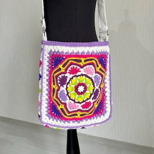 以下が含まれることがあります： 白のクロシェのショルダーバッグ。ピンク、パープル、イエロー、グリーンのカラフルな花柄のデザイン。バッグには白いストラップが付いています。