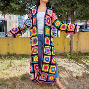 May include: A long, black crochet cardigan with a colorful granny square pattern. The cardigan has a deep V-neck and long sleeves. The squares are in shades of pink, yellow, blue, green, and orange.