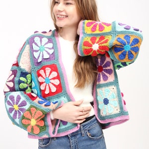 May include: A colourful crocheted cardigan with a floral pattern. The cardigan has a pink trim and is made up of squares with different coloured flowers.