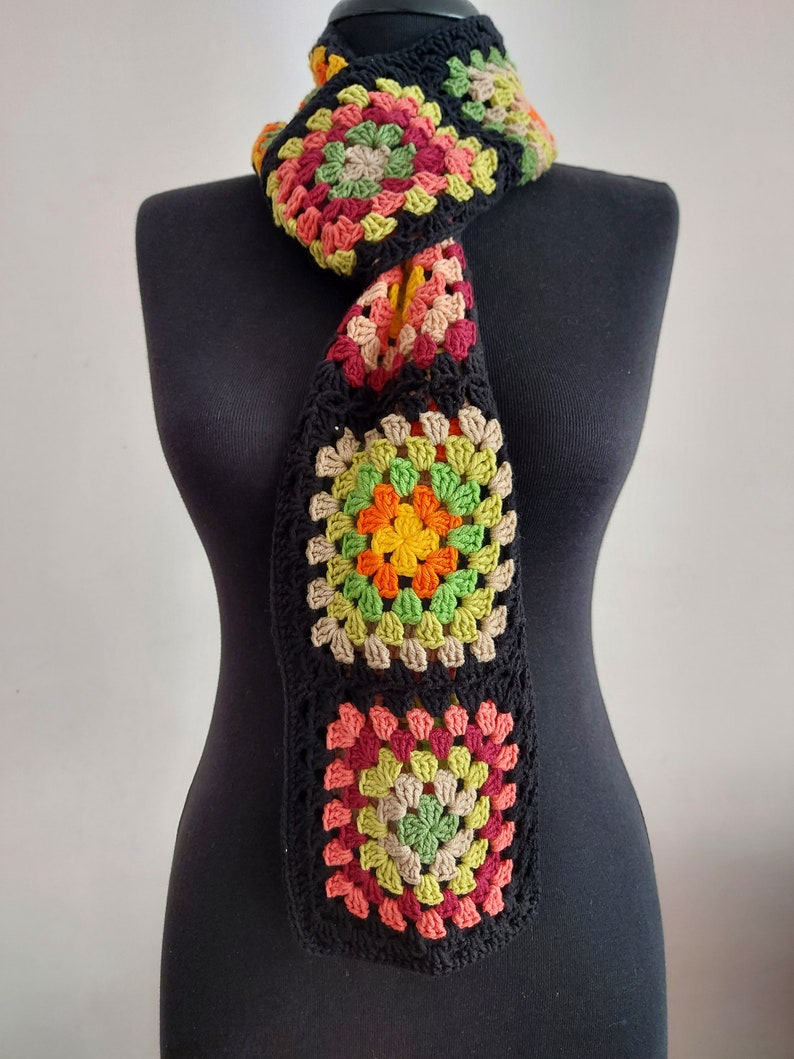 May include: A crocheted scarf with a black background and colorful granny squares in shades of red, orange, yellow, green, and brown.