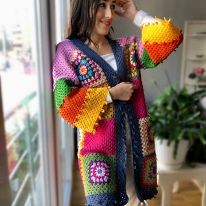 Könnte beinhalten: Eine bunte gehäkelte Strickjacke mit Patchwork-Muster aus Quadraten in den Farben Rosa, Grün, Orange, Gelb und Blau. Die Strickjacke hat einen V-Ausschnitt und lange Ärmel.