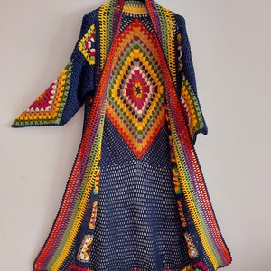 May include: A long, navy blue crochet cardigan with a colorful diamond pattern in the center and rainbow stripes along the sides. The cardigan has a wide, open neckline and long sleeves.