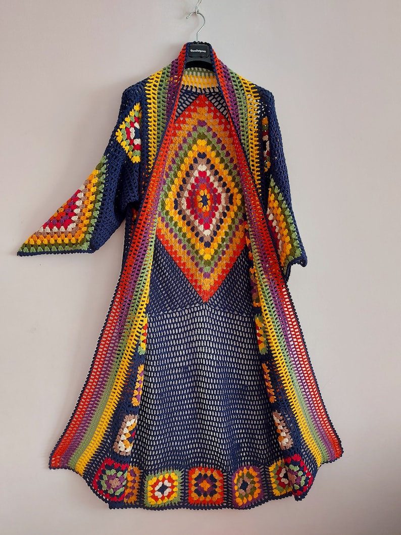 May include: A long, navy blue crochet cardigan with a colorful diamond pattern in the center and rainbow stripes along the sides. The cardigan has a wide, open neckline and long sleeves.