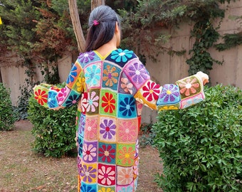 May include: A long, colourful crochet cardigan with a floral pattern. The cardigan is made up of squares with different coloured flowers in each square. The cardigan is being worn by a person standing in a garden.