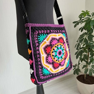 May include: A black crocheted shoulder bag with a colorful floral design. The bag has a purple trim and a black adjustable strap.