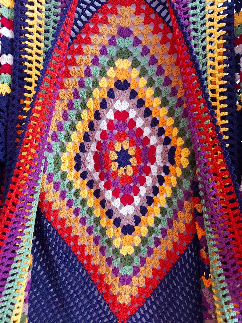 May include: A colorful crocheted shawl with a diamond pattern in shades of red, orange, yellow, green, blue, purple, and white. The shawl is made with a granny square stitch and has a navy blue border.