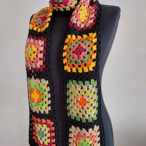 May include: A crocheted scarf with a black background and colorful granny squares. The squares are made in shades of green, orange, yellow, pink, and brown.