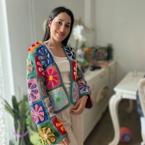 May include: A long-sleeved cardigan made of multi-colored crochet granny squares featuring various flower motifs in shades of blue, pink, red, green, and yellow. The garment has an open front design and is worn over a simple white tank top.