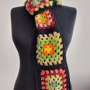 May include: A crocheted scarf with a black background and colorful granny squares in shades of red, orange, yellow, green, and brown.