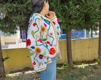 Op de afbeelding: Een wit gebreid vest met kleurrijke bloemenborduursels. Het vest heeft lange mouwen en een ritssluiting.