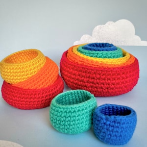 Puede incluir: Un conjunto de cuencos anidables de crochet de color arcoíris. Los cuencos están hechos de hilo en tonos de rojo, naranja, amarillo, verde y azul. Los cuencos están apilados uno encima del otro, con el cuenco más grande en la parte inferior y el más pequeño en la parte superior.