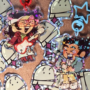May include: Two colorful character charms with glitter accents. The first charm features a cartoon character with long black hair, wearing sunglasses and a red jacket, playing a guitar. The second charm features a cartoon character with short black hair, wearing a blue jacket and holding a small object. Both charms are attached to a blue star-shaped keychain.