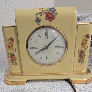 May include: A vintage, cream-colored ceramic clock with a gold-tone bezel and accents. The clock face has black Roman numerals and hands. Floral designs in red, blue, and yellow adorn the clock's face and sides. The clock has a rectangular shape.