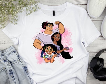 May include: White t-shirt with a pink watercolor background featuring a cartoon illustration of three women. The women are standing together and smiling. The woman on the left is wearing a purple shirt and has her arm raised in the air. The woman in the middle is wearing a pink dress and has her arm around the woman on the right. The woman on the right is wearing a blue dress and has her hand on her hip.