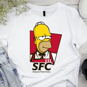 Pode incluir: Camiseta branca com uma imagem de desenho animado do Homer Simpson vestindo um uniforme vermelho e amarelo com o texto "SFC Simpsons Fried Chicken".