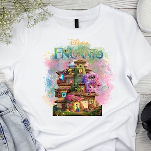 May include: White t-shirt featuring the Disney Encanto logo and a colorful illustration of the Madrigal family's house. The design includes the word "Encanto" in a vibrant, multi-colored font, with a watercolor-style background.